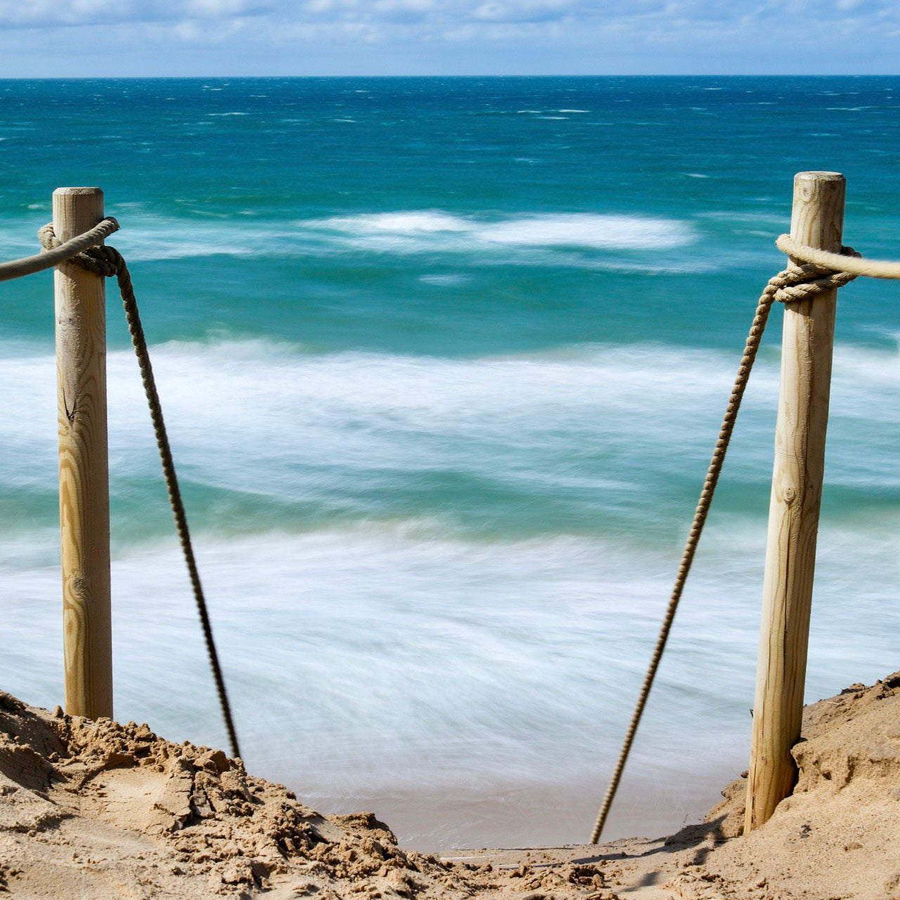 Das rd-pictures Motiv "Abstieg" zeigt einen extrem steilen Abstieg von einer Dünen herunter zum Meer. Links und rechts sieht man Holzpfähle, die mit einem Tau umwickelt sind. Im Hintergrund sieht man die tosende Nordsee.