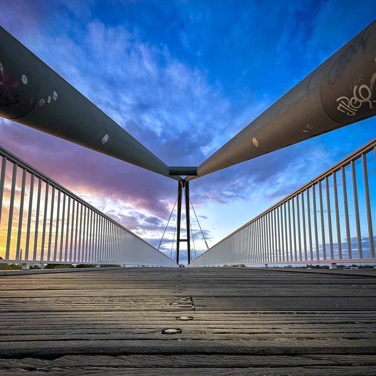 Besondere Perspektive der Fußgängerbrücke im Düsseldorfer MedienHafen. Das Foto wurde auf Stehhöhe aufgenommen. Im Vordergrund sieht man Holzplanken, seitlich ein typisches Gittergeländer, welches perspektivisch zusammenläuft. Der Himmel ist in blau getaucht. Einige Wolken werden von der Sonne in gelb und lila gefärbt.