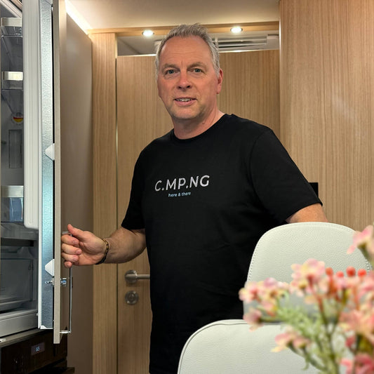 Mann trägt ein schwarzes Camping T-Shirt aus Biobaumwolle mit stylishen Aufdruck in grau und türkis. Untereinander stehen dort C.MP.NG und darunter here & there in used-Optik. Im Hintergrund sieht man die Küxhe eines Wohnmobils.