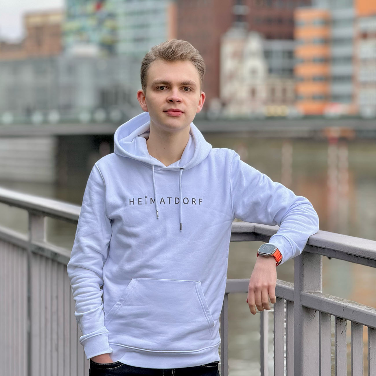 Junger Mann mit Düsseldorf Heimatdorf Hoodie in weiß im MedienHafen. Auf der Brust ist in grau der Schriftzug Heimatdorf aufgedruckt.