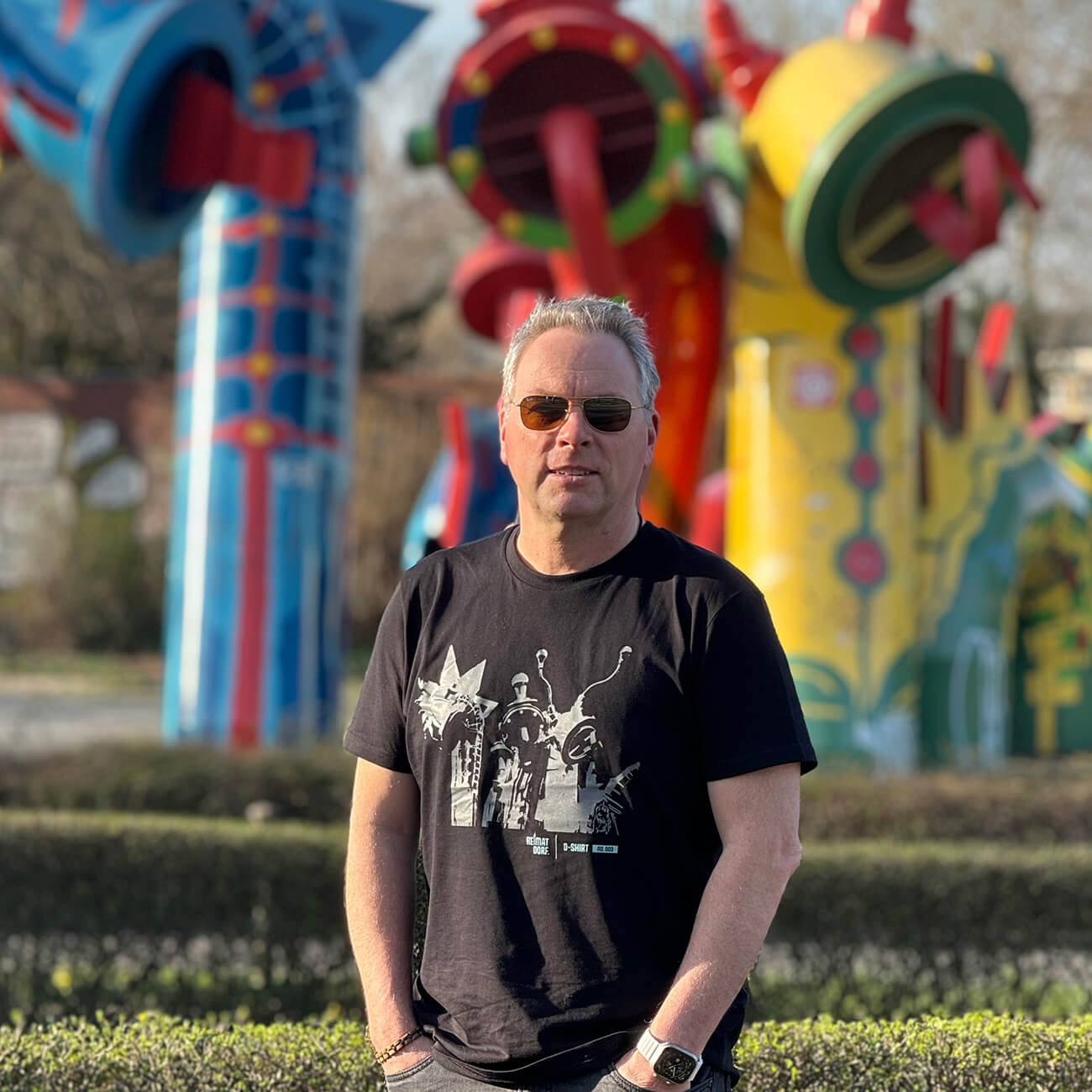 Mann steht mit schwarzem Düsseldorf T-Shirt vor Nessie Familie in Düsseldorf Bilk. Auf dem T-Shirt ist in grau die Drachenfamilie als Grafik aufgedruckt.