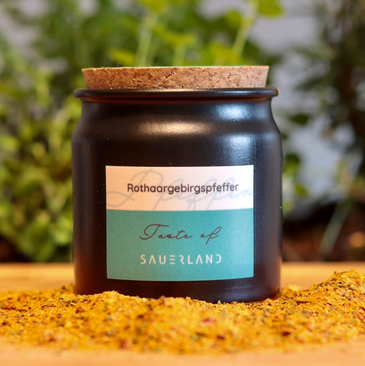 Dunkelgraues Gewürztöpfchen mit Korkenverschluss steht auf einem Holzuntergrund mit Rothaargebirgspfeffer. Auf dem Töpfchen befindet sich ein Aufkleber in weiß-türkis mit dunkelgrauem Aufschrift: Rothaargebirgspfeffer, Taste of Sauerland.