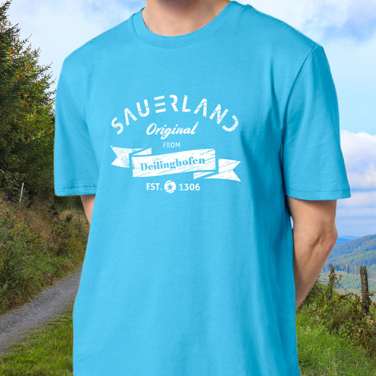 Mann in azurblauem T-Shirt mit weißem Aufdruck Sauerland Original from Deilinghofen Established 1306 steht auf einem Weg in Sauerländer Natur