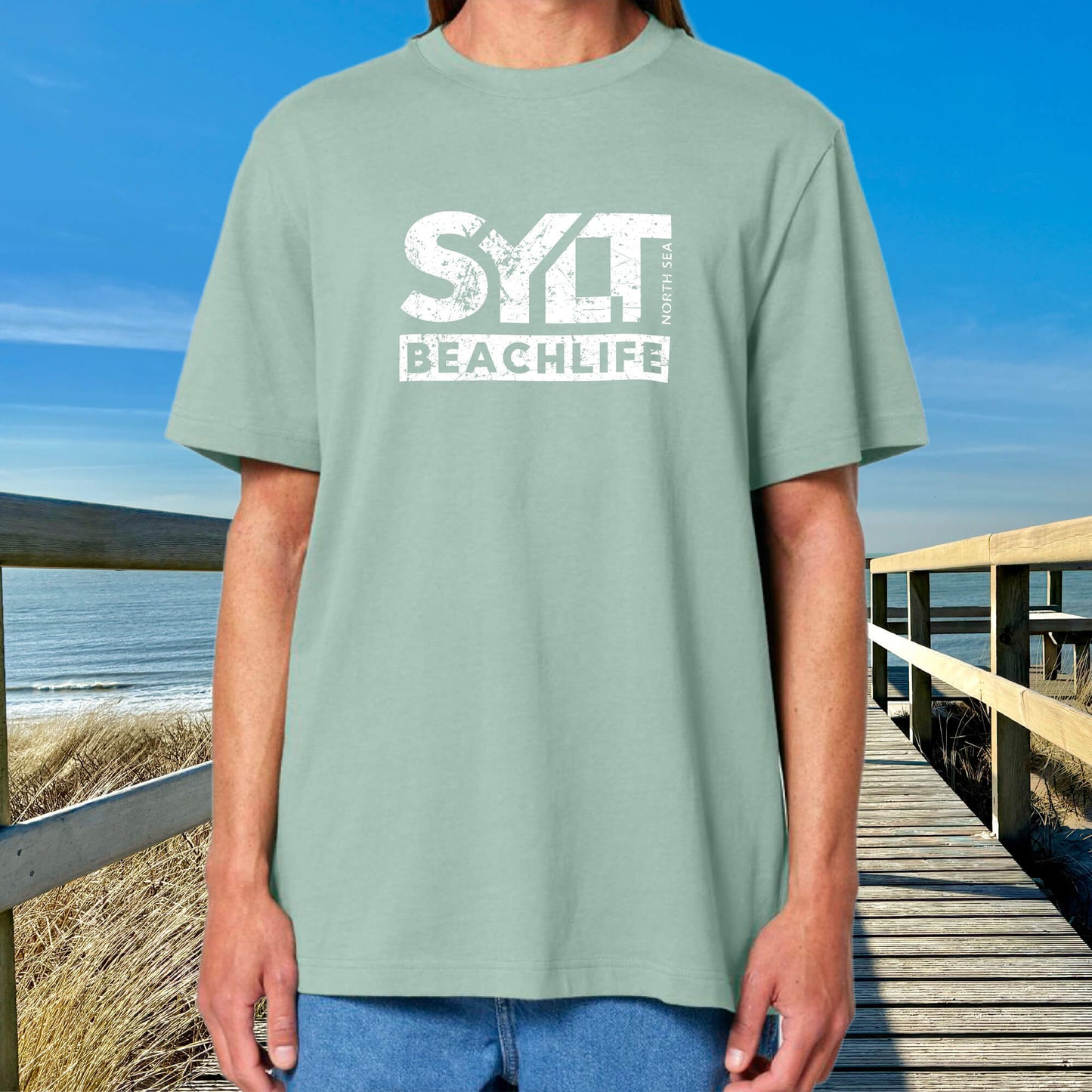 Mann in heidegrünem Sylt T-Shirt steht auf Holzsteg mit Meerblick und Dünenlandschaft im Hintergrund