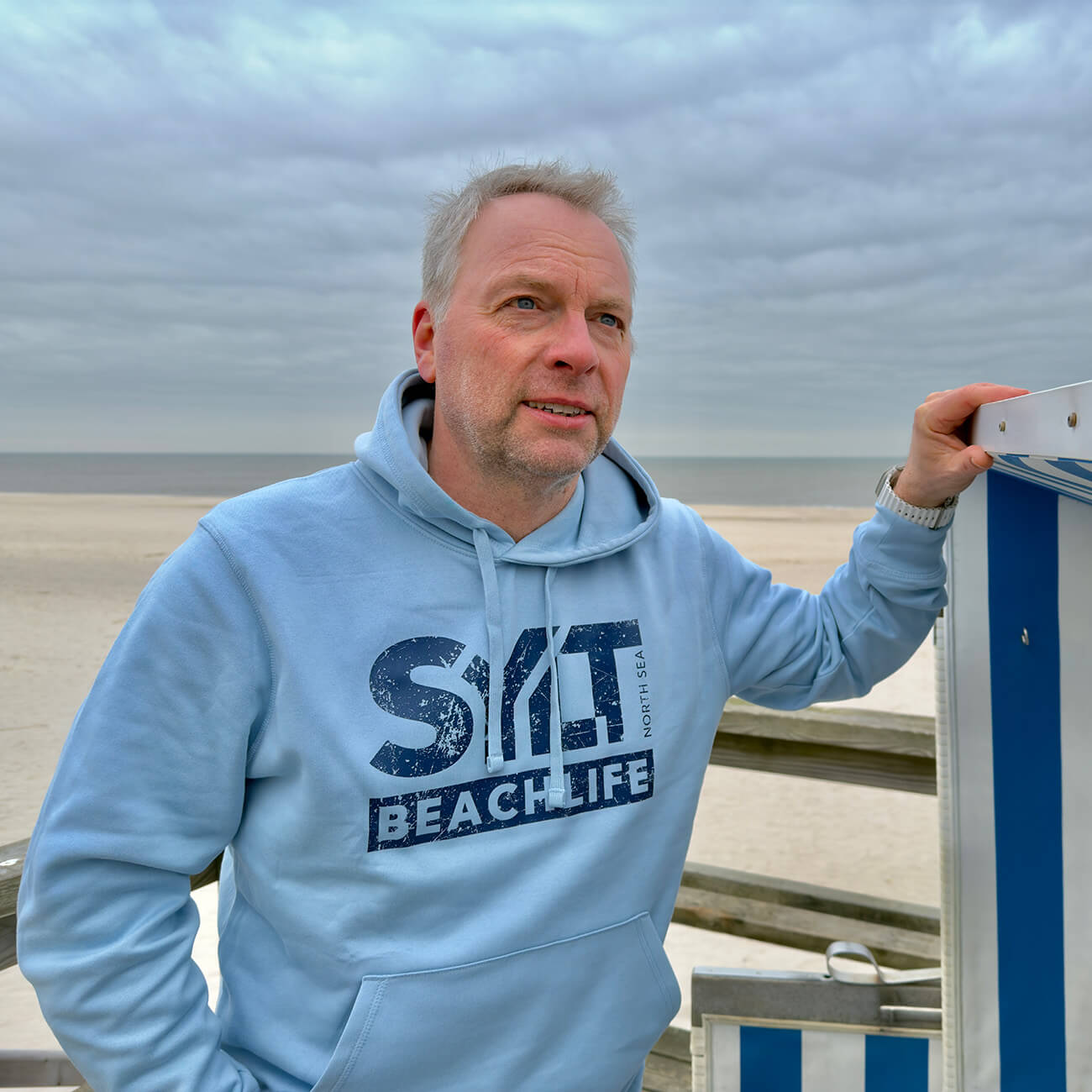 Mann mit hellblauem Sylt Hoodie und dunkelblauem Aufdruck "Sylt Beachlife" steht an einen Strandkorb gelehnt. Im Hintergrund ist der Strand und die Sylter Nordsee zu sehen.