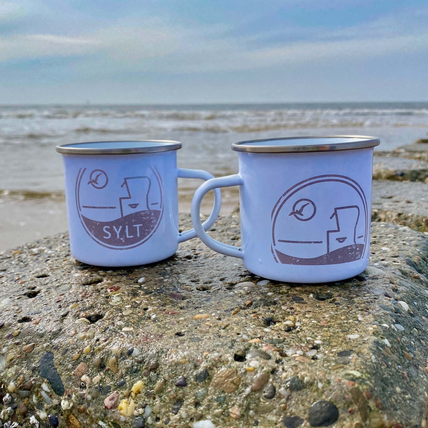 Zwei Sylter Emaillebecher mit silbernem Rand stehen auf einer Buhne. Im Hintergrund ist das Meer zu sehen. Auf den weißen Emailletassen ist eine runde Grafik aufgedruckt, die einen Sylter Nordseestrandkorb, eine Möwe die durch die Sonne fliegt sowie der Begriff Sylt zeigt.