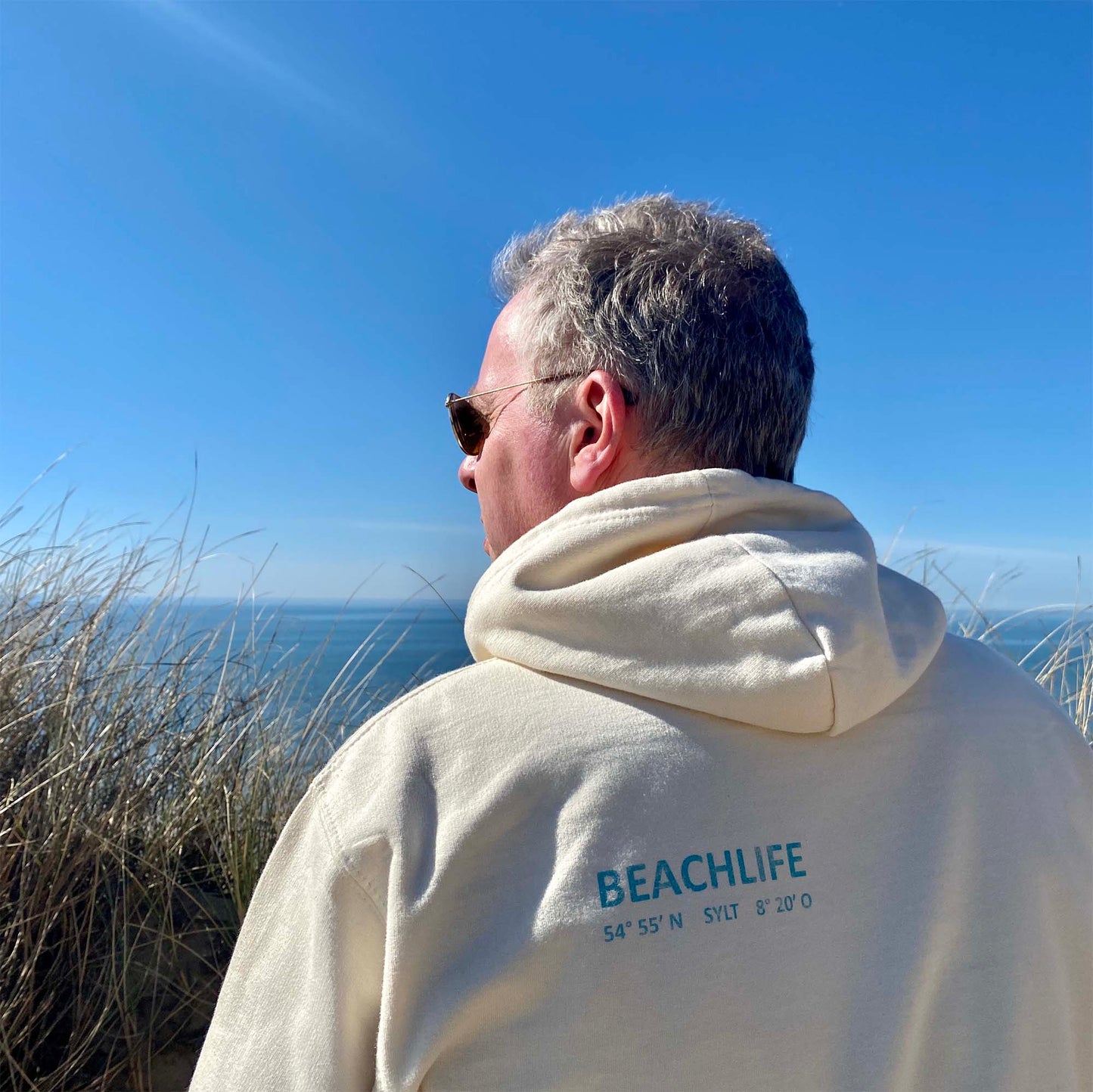 Mann mit beidem Hoodie ist von hinten zu sehen, während er den Ausblick auf Sylt genießt. Im Hintergrund sieht man Dünen und das Meer. Auf der linken Schulter unter der Kapuze ist in türkis das Wort Beachlife sowie die Koordinaten von Sylt sowie der begriff Sylt aufgedruckt.