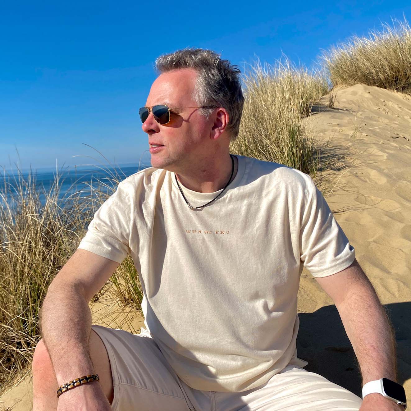 Mann sitzt in einem beigen T-Shirt in den Dünen von Sylt. Auf dem beigen T-Shirt ist oben mittig in klein ein Aufdruck mit den Koordinaten von Sylt zu finden. Die Farbe des Aufdrucks ist Kupfer.