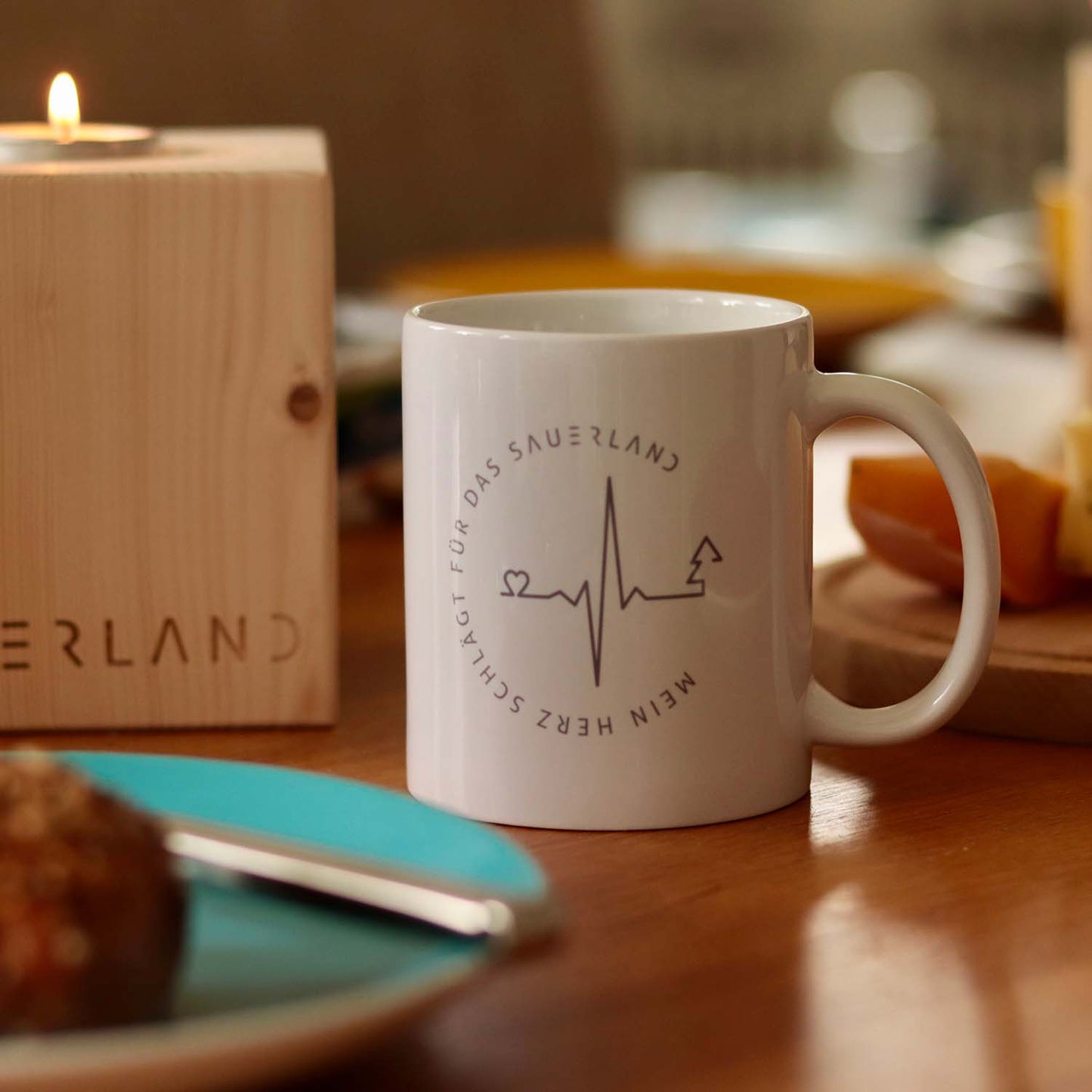 Weißer Becher mit grauem Aufdruck. Das Motiv zeigt einen Herzschlag der in einen Tannenbaum mündet und wird vom Spruch mein Herz schlägt für das Sauerland umrundet. Der Becher steht auf einem gedeckten Frühstückstisch. Im Anschnitt ist der Sauerland Teelichtwürfel zu sehen.