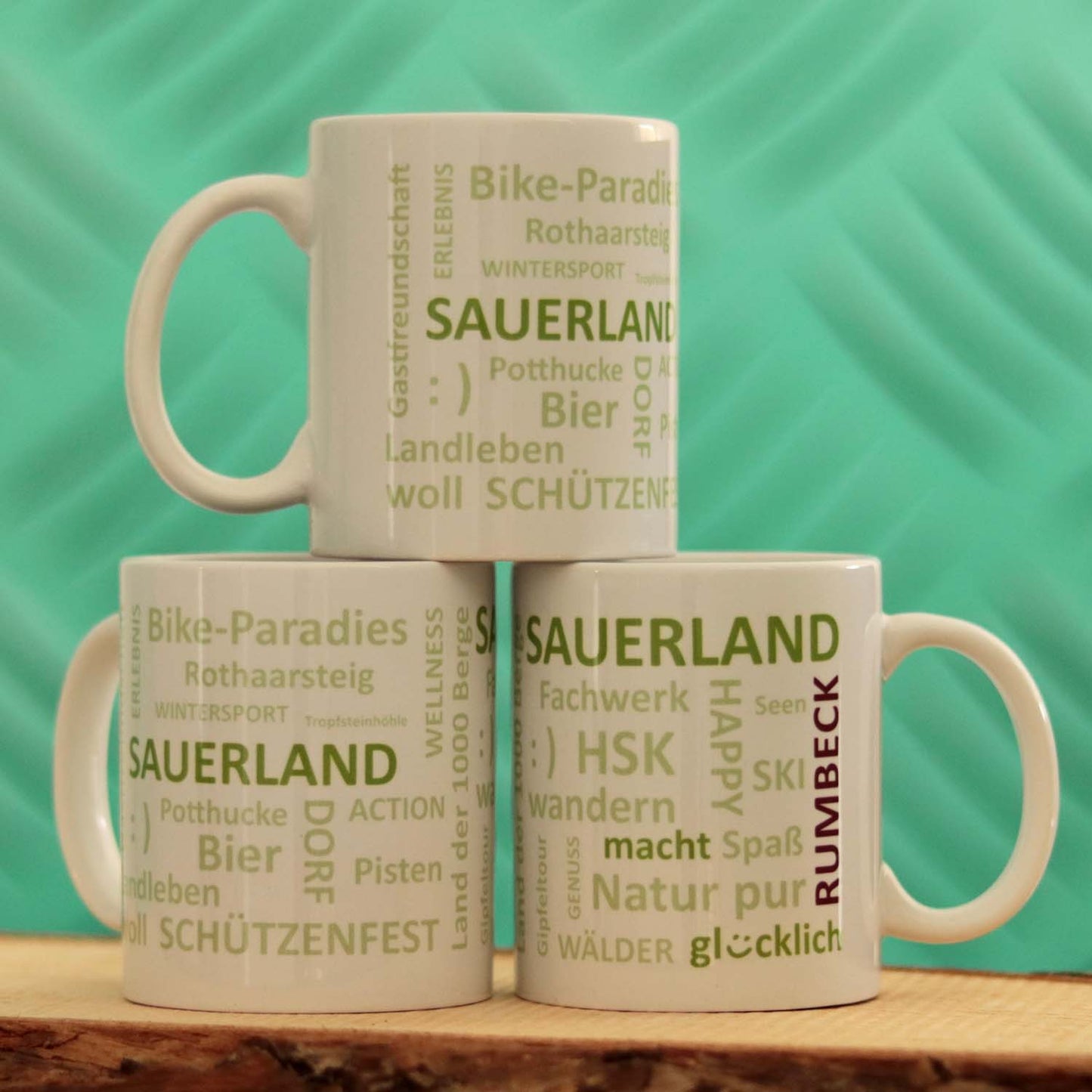 Drei weiße Keramik-Becher stehen in Pyramidenform aufeinandergestapelt auf einem Holzbrett. Die Becher sind mit typischen Begriffen aus dem Sauerland in grün bedruckt. Dies sind u.a. Schützenfest, soll, Landleben, Sauerland macht glücklich. Links neben dem Henkel ist der Dorfname Rumbeck in dunkelrot aufgedruckt. Die Dorfbecher kann man mit jedem beliebigen Dorfnamen produzieren lassen.