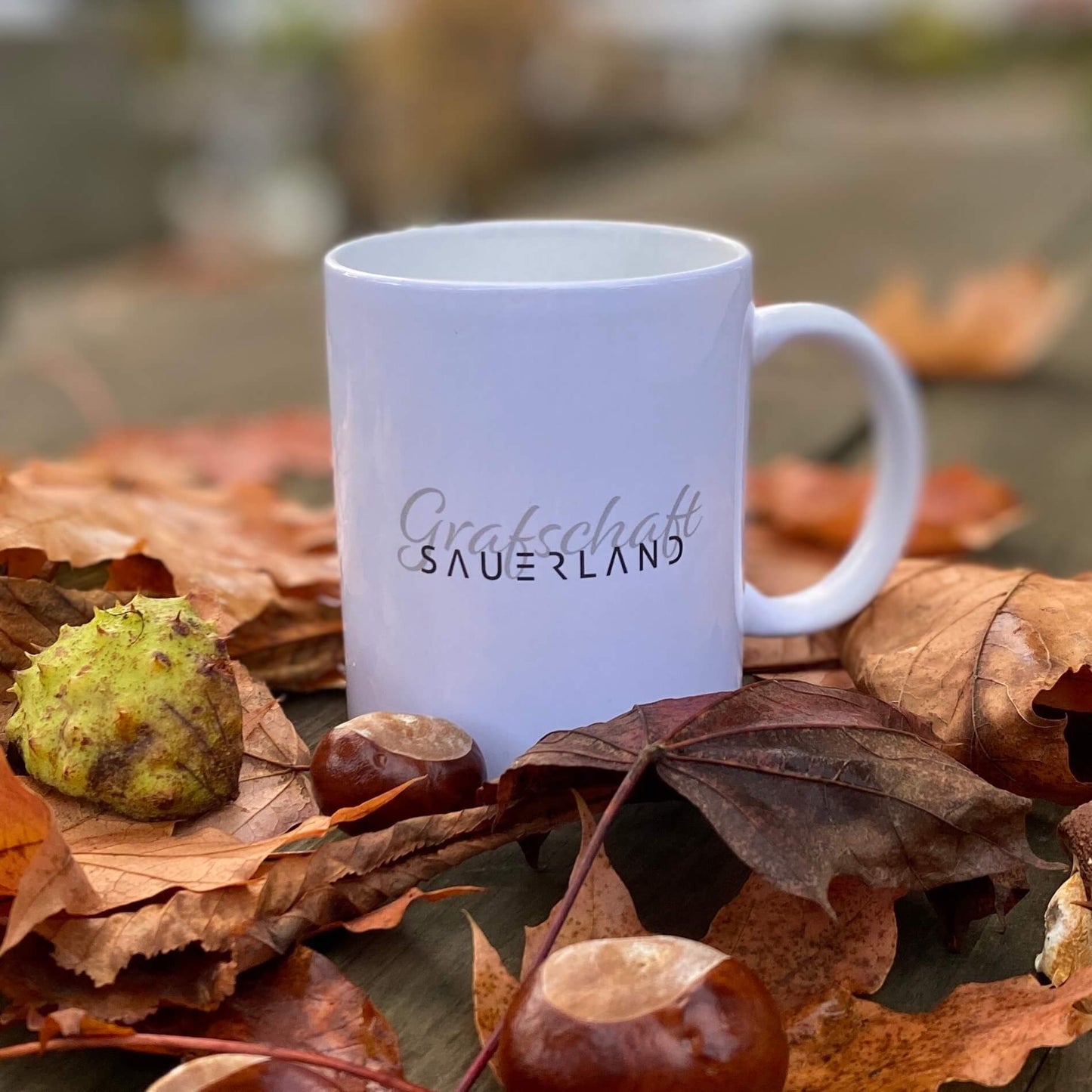 Weiße Kaffee- oder Tee-Tasse mit dem Dorfnamen Grafschaft in geschwungener Schrift sowie mit dem New Sauerland Schriftzug beschriftet. Der Becher steht auf einem Holztisch mit herbstlichem Laub und Kastanien.
