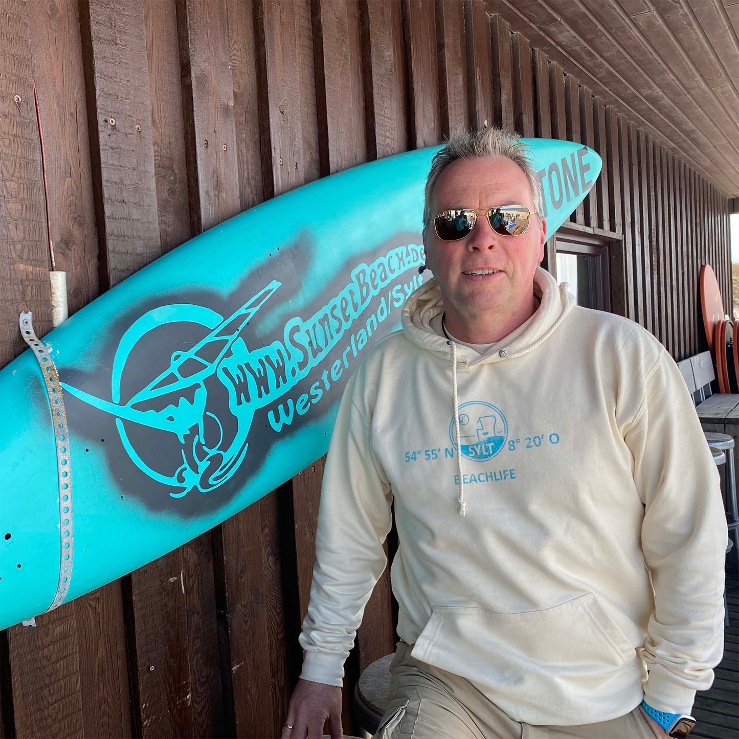 Mann steht mit Hellbeige Sylt-Hoodie vor einem Türkisen Surfbrett. Der Hoodie ist vorne in türkis mit den Koordinaten von Sylt sowie einem Strandkorb bedruckt. Darunter ist der Begriff Beschlief aufgedruckt.
