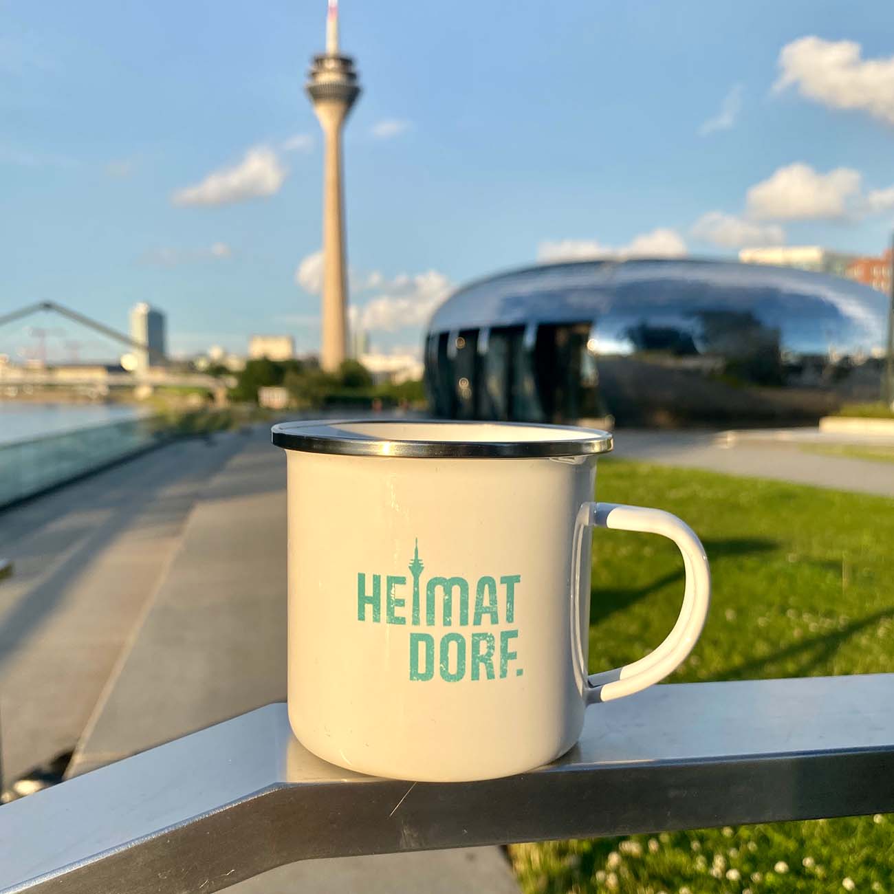 Weißer Emaillebecher mit silbernem Rand und Türkisen HeimatDorf Logo von Düsseldorf steht auf einem Geländer im MedienHafen mit Blick auf den Rheinturm.