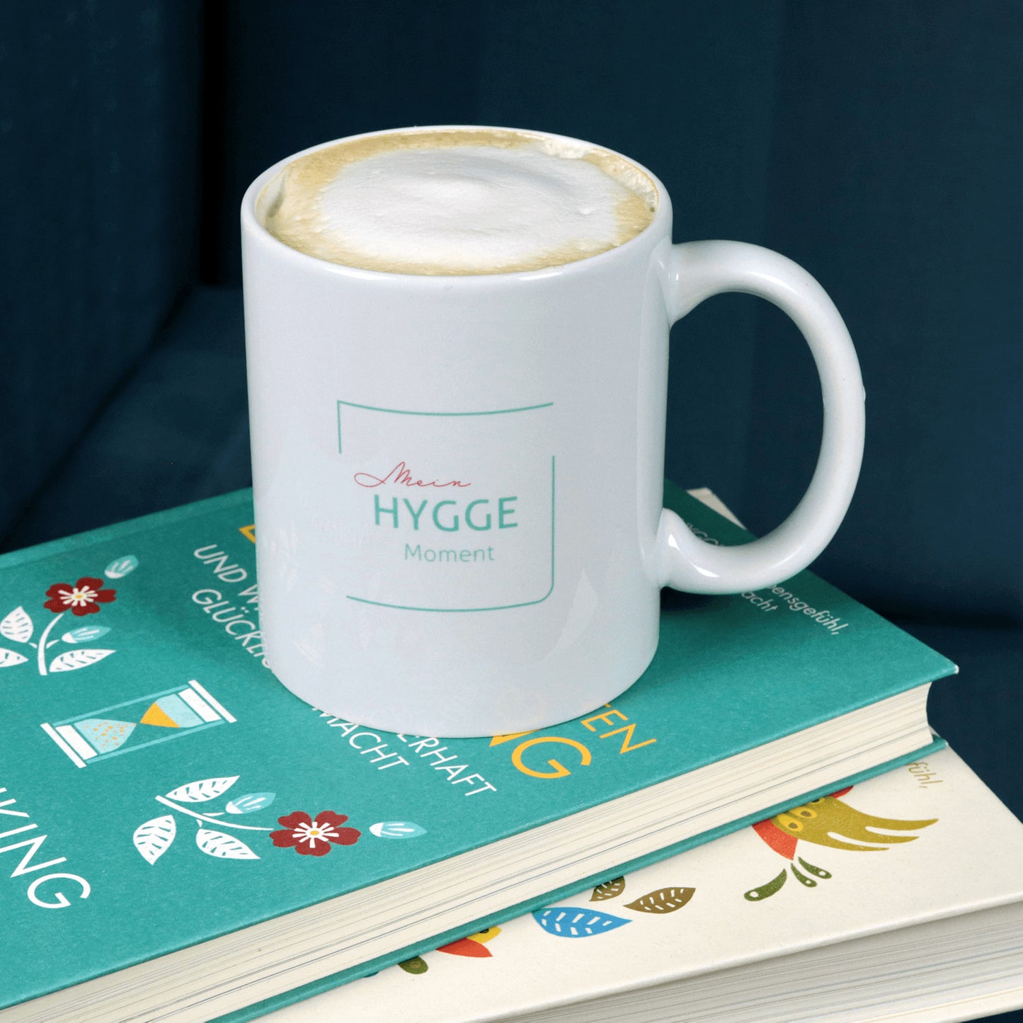Weißer Keramik-Becher mit Aufdruck Mein Hygge Moment steht auf zwei Büchern.