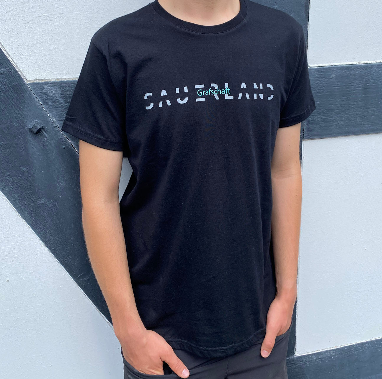 Mann steht mit einem schwarzen Sauerland T-Shirt vor einem Fachwerkhaus. Das schwarze T-Shirt ist imit einem grauen SAUERLAND Schriftzug versehen, der in der Mitte eine schwarze Linie als Aussparung hat. Mittig in der Aussparung ist in türkis der Dorfname Grafschaft eingedruckt.