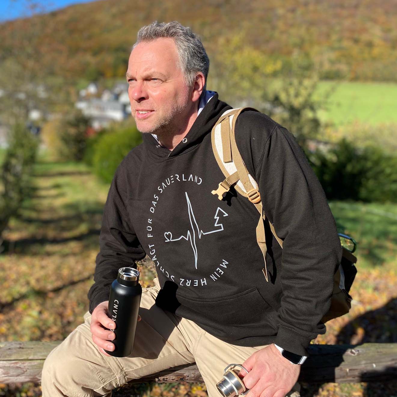 Mann sitzt mit schwarzem Hoodie und Rucksack im Sauerland auf einem Holzbalken. Auf dem Hoodie ist in grau und zeigt einen Herzschlag der mit der Grafik Herz anfängt und mit einer grafik von einem Tannenbaum aufhört. Drumherum ist in rund geschrieben: Mein Herz schlägt für das Sauerland.