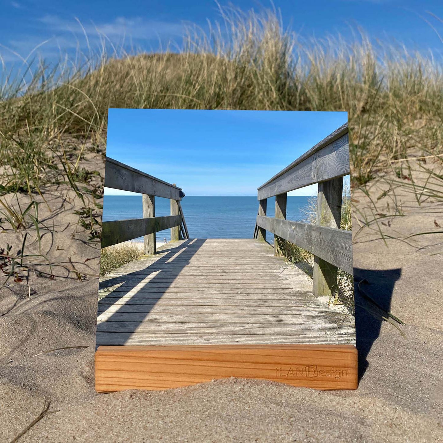 Ein leuchtendes Acrylbild steht in einem hölzernen Standbilderhalter in den Sylter Dünen. Auf dem Bild ist ein Holzsteg mit Blick auf die Nordsee zu sehen.