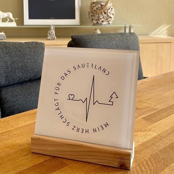 Hellgraues Acrylbild mit dunkelgrauem Logoaufdruck "Mein Herz schlägt für das Sauerland". Der Schriftzug ist in rund aufgedruckt. In der Mitte befindet sich eine Herzschlaggrafik, die mit einem herz startet und in einem Tannenbaum endet.
