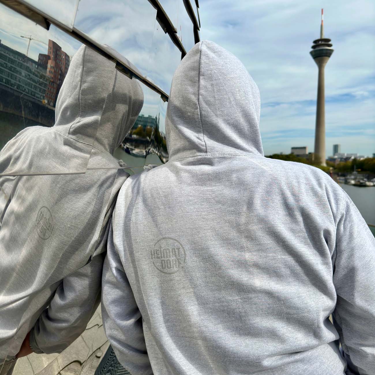 Mann ist mit einem hellgrauen Hoodie von hinten zusehen. Er steht angelehnt an das Pebbels im Düsseldorfer MedienHafen. Auf der linken Schulter des Hoodies ist das HeimatDorf logo in rund aufgedruckt. Im Hintergrund ist der Düsseldorfer Rheinturm zu sehen, der auch Inhalt des HeimatDorf Logos ist.