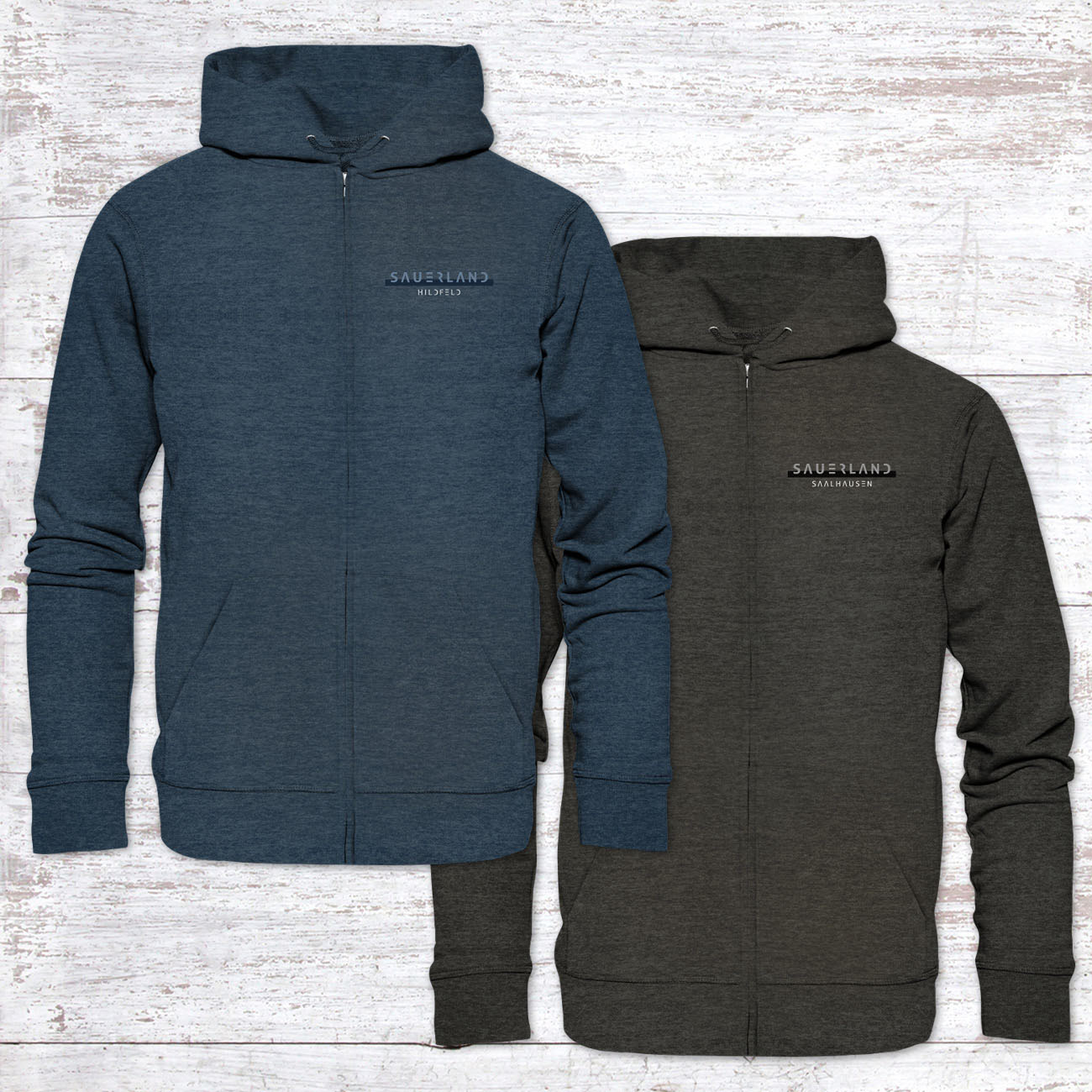 Zwei Hoodie Zipperlein-Jacken in dunkelblau und dunkelgrau liegen auf einem Holzhintergrund. Jeweils auf der linken Brust ist der new Sauerland Schriftzug aufgedruckt. Darunter in weiß wird der Wunschdorfname eingedruckt.