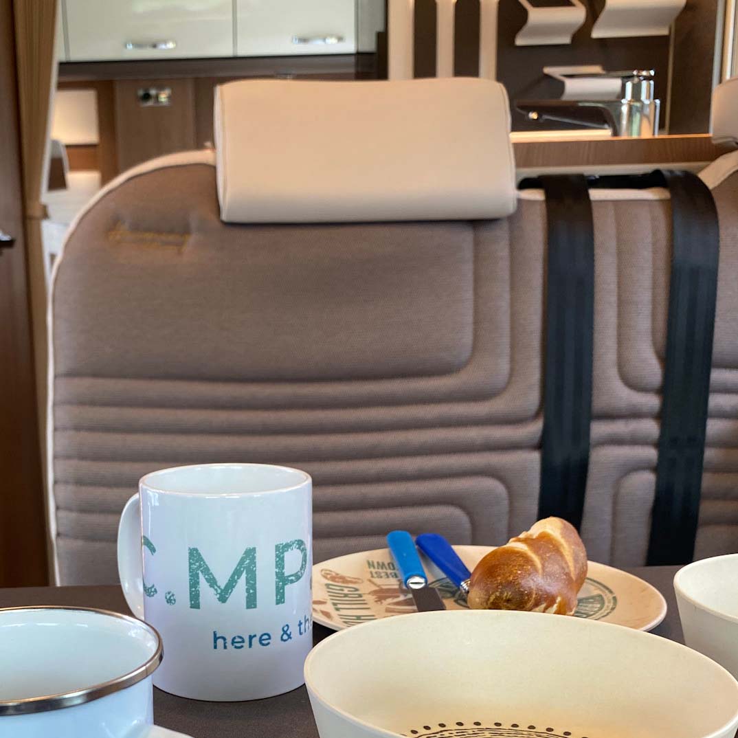 Camping-Becher aus weißer Keramik mit großem C.MP.NG here and there Aufdruck auf einem gedeckten Tisch im Wohnmobil