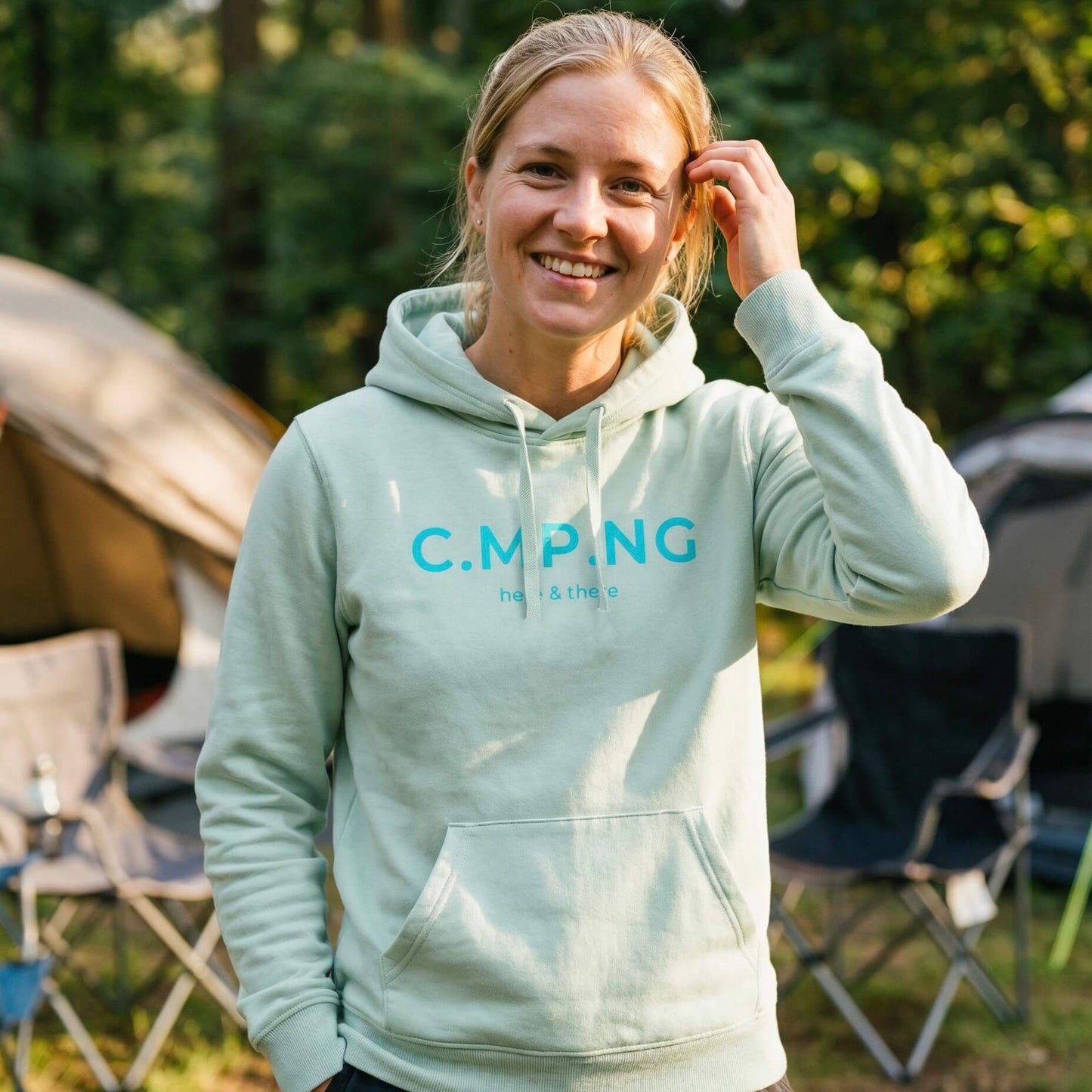 Eine blonde Frau trägt beim camping ihren C.MP.NG original Hoodie in Aloe.