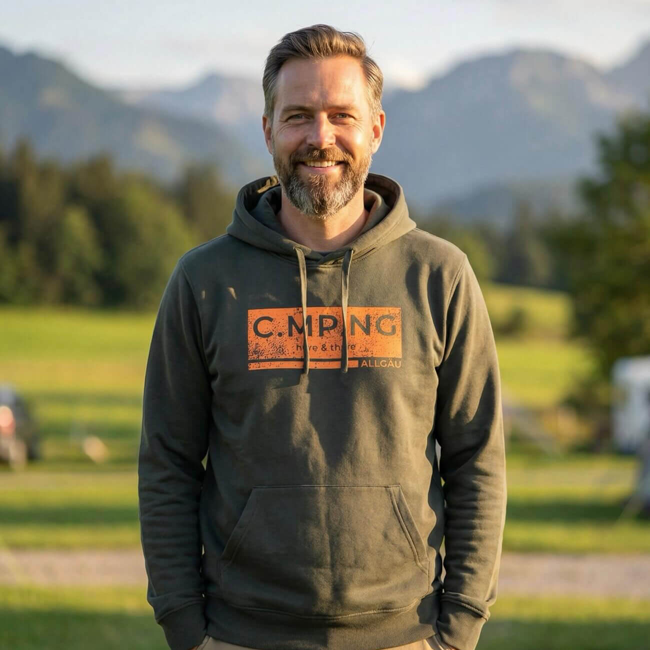 Ein mittelalter Mann steht vor einem bergischen Hintergrund mit einem dunkelgrünen Hoodie von C.MP.NG an