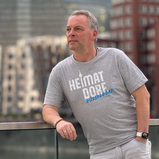 Mann steht mit Düsseldorf T-Shirt im Medienhafen vor den Gehry-Bauten. Das T-Shirt ist heather grey und in weiß mit dem HeimatDorf-Logo im Graffiti-Look bedruckt. Schräg darunter prangt der Name Düsseldorf in auffälligem blau.