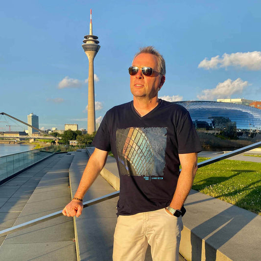 D-Shirt No. 002 – Das Düsseldorf T-Shirt.Mann steht im Düsseldorfer MedienHafen vor dem Rheinturm. Im Hintergrund sieht man die Stadt Düsseldorf. Der Mann hat ein dunkelblaues T-Shirt mit V-Ausschnitt an. Auf dem T-Shirt ist ein großer Aufdruck mit den schimmernden Scheiben des Rheinturms zu sehen. 