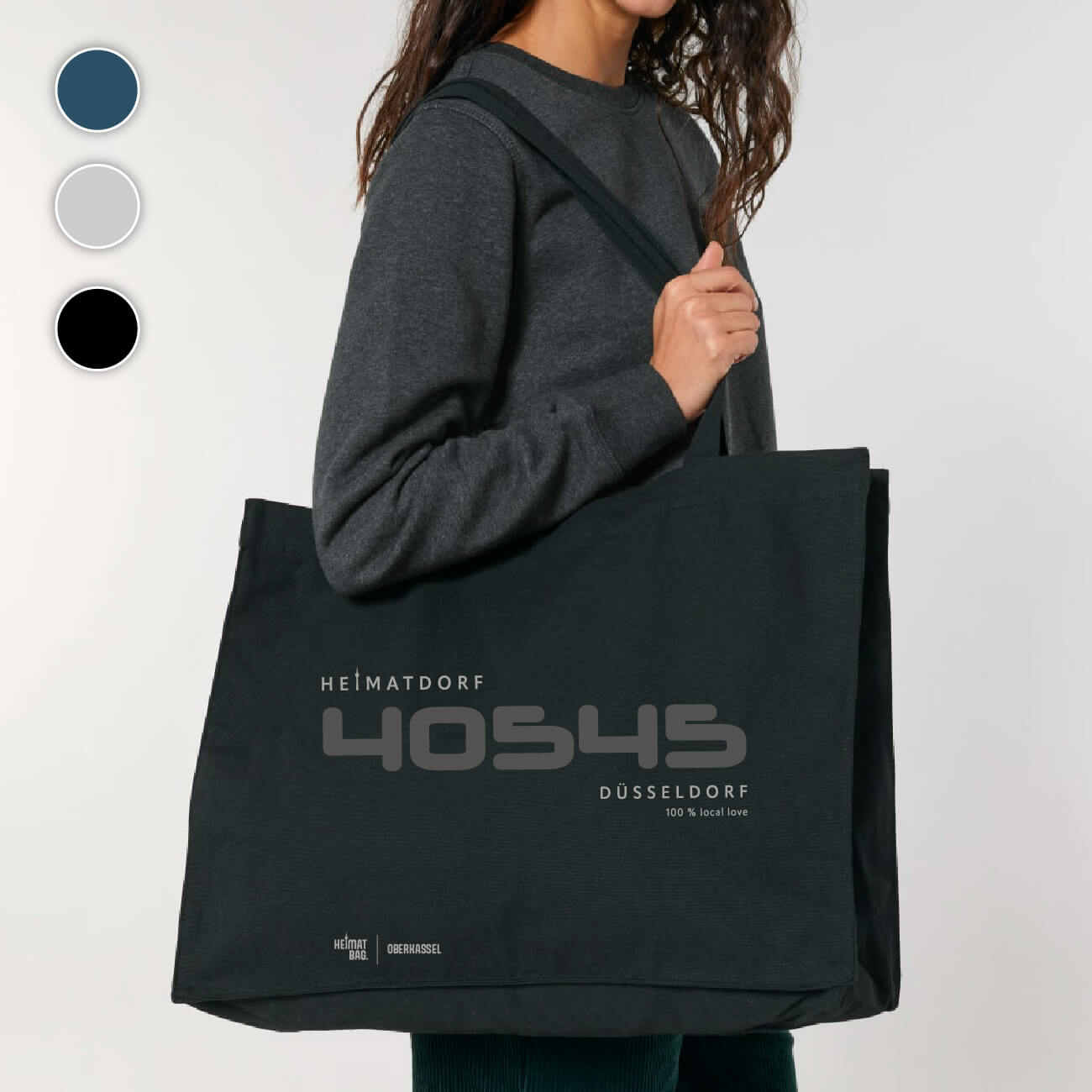 Frau steht mit schwarzer Shopping-Tasche über der Schulter vor einem grauen Hintergrund. Groß ist in dunkelgrau die PLZ 40545 aufgedruckt. Darüber in hellgrau Heimatdorf und darunter Düsseldorf 100 % local love. Am unteren Saum befindet sich ein hellgrauer Aufdruck Heimat-Bag Oberkassel.