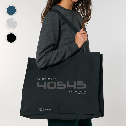 Frau steht mit schwarzer Shopping-Tasche über der Schulter vor einem grauen Hintergrund. Groß ist in dunkelgrau die PLZ 40545 aufgedruckt. Darüber in hellgrau Heimatdorf und darunter Düsseldorf 100 % local love. Am unteren Saum befindet sich ein hellgrauer Aufdruck Heimat-Bag Oberkassel.