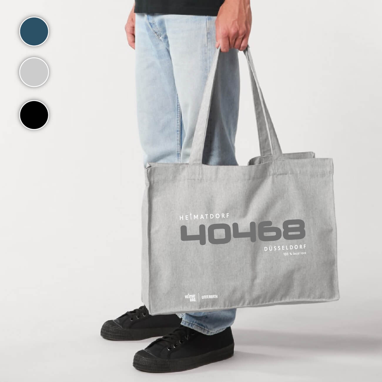 Mann hält eine hellgraue Düsseldorf-Tasche in der Hand. Auf der Tasche ist ein zweifarbiger Aufdruck platziert. In der Mitte steht in grau die Postleitzahl 40468 und darüber in weiß der Begriff Heimatdorf. Unter der Postleitzahl steht in weiß Düsseldorf 100 % local love. Unten links kurz über dem Saum befindet sich ein weißer Aufdruck Heimat-Bag Unterrath