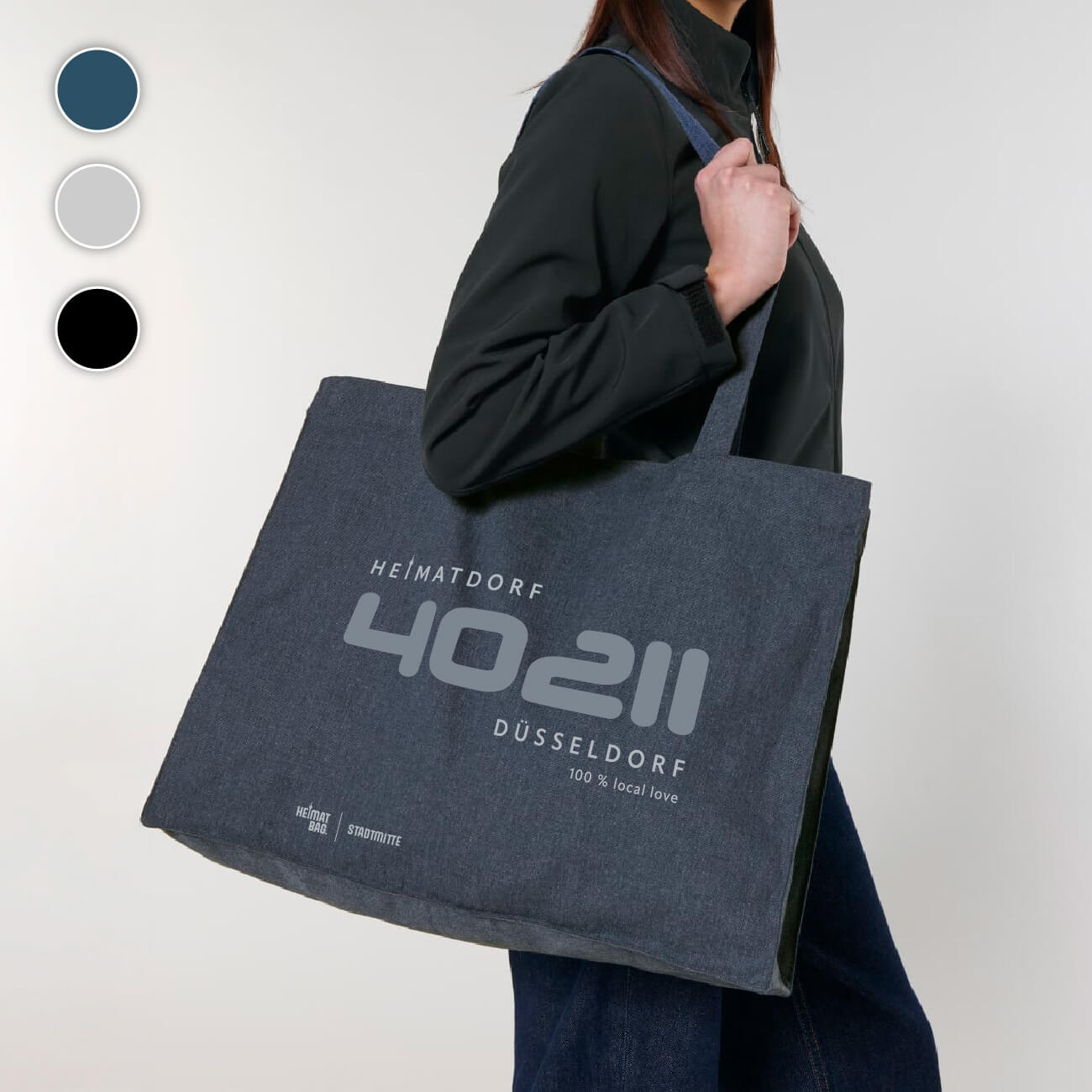 Eine Frau trägt eine Baumwolltasche in navy-blau über der Schulter.  In mittelblau ist die Postleitzahl 40211 aufgedruckt. Darunter in hellgrau Düsseldorf 100 % local love. Darüber ist das Heimatdorf Logo mit stilisiertem Rheinturm als i aufgedruckt. Links unten über dem Saum ist in hellgrau der Begriff Heimat-Bag Stadtmitte aufgedruckt.