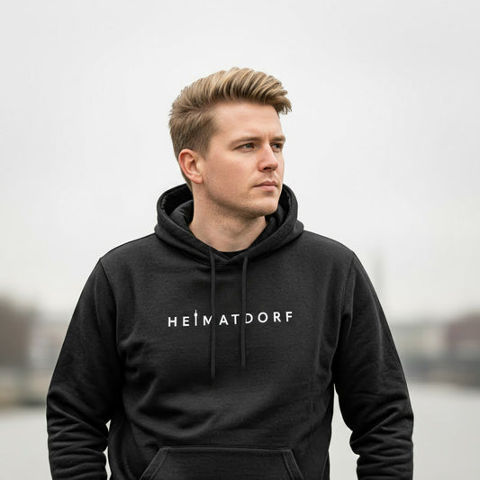 Mann hat schwarzen Heimatdorf Düsseldorf Hoodie an. Auf der Brust ist in weiß der Schriftzug Heimatdorf aufgedruckt. Das i ist durch die Silhouette des Rheinturms ersetzt.