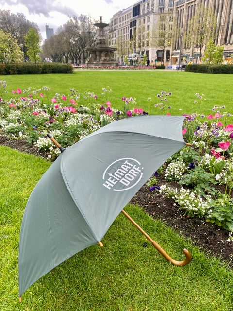 Grauer HeimatDorf Schirm liegt auf einer Wiese mit Blumenbeet auf der Königsallee in Düsseldorf. Auf einem Segment des Schirms ist das HeimatDorf-logo in weiß aufgedruckt.