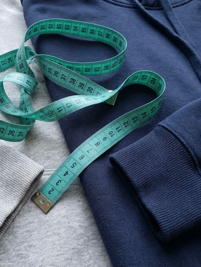 Ein grauer und ein blauer Hoodie liegen gefaltet, darauf ein maßband