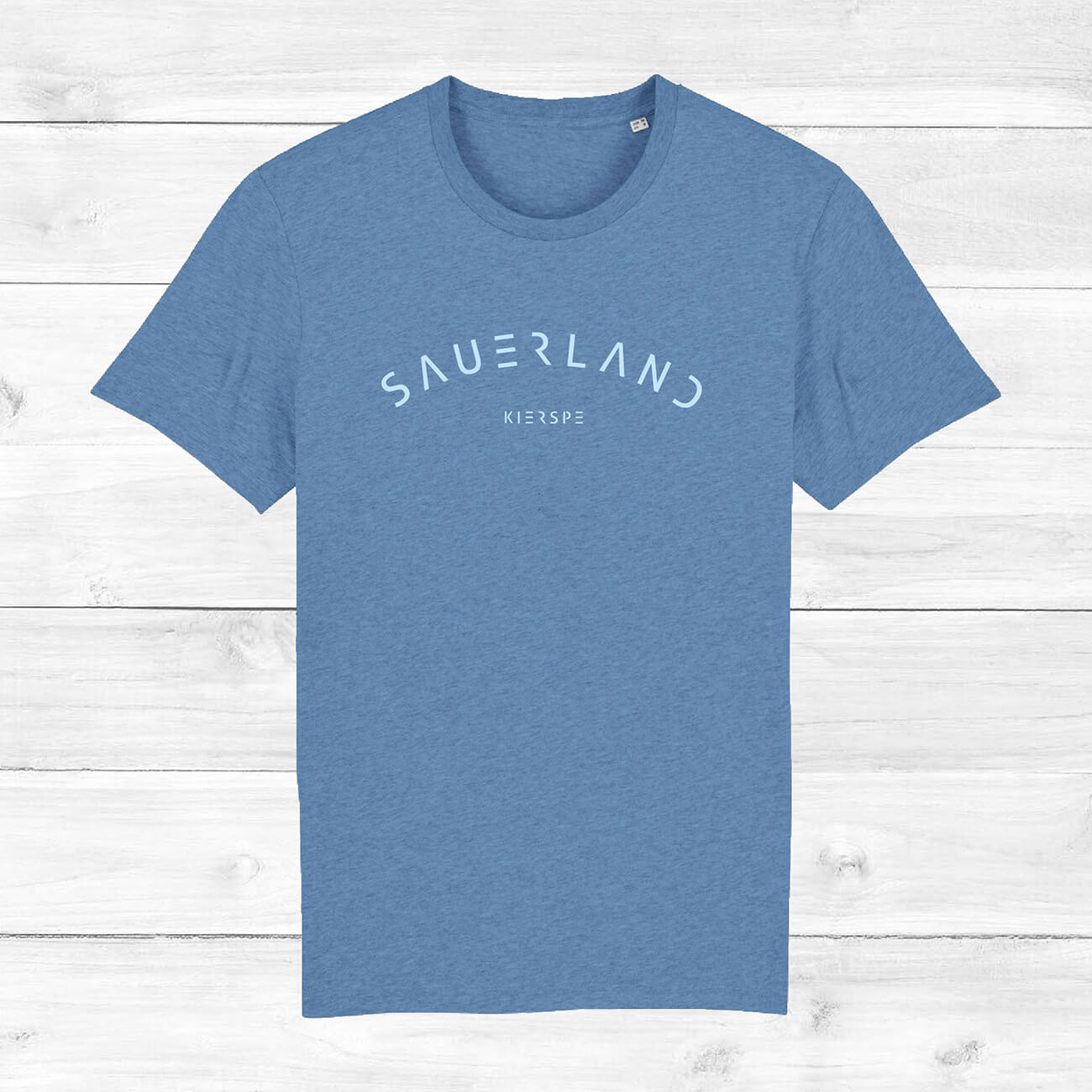 Mittelblaues T-Shirt mit leicht gerundetem Sauerland-Schriftzug liegt auf einem Holzhintergrund. In der Mitte unter dem Sauerland-Schriftzug ist der Dorfname Kierspe eingedruckt. Der Aufdruck ist hellblau.