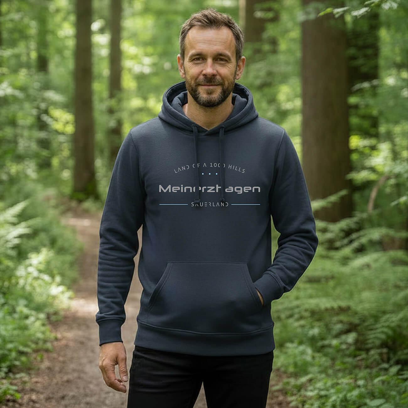 Ein Mann welcher im Wald steht mit einem blau-grauen Hoodie, Aufdruck des Hoodies ist: Land of a 1000 Hills - Meinerzhagen - Sauerland