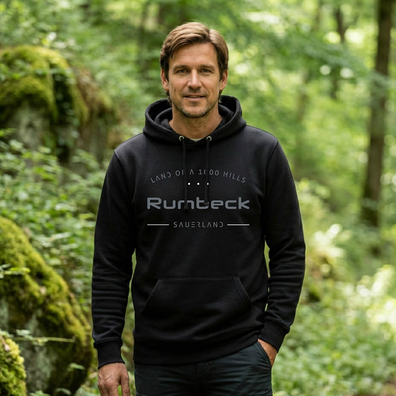 Ein Mann welcher im Wald steht mit einem schwarzen Hoodie, Aufdruck des Hoodies ist: Land of a 1000 Hills - Rumbeck - Sauerland