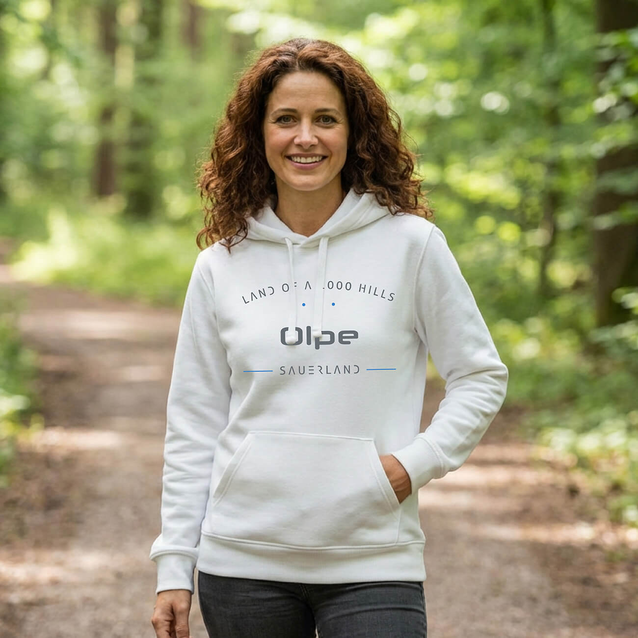 Eine Frau welche im Wald steht mit einem weißen Hoodie, Aufdruck des Hoodies ist: Land of a 1000 Hills - Olpe - Sauerland