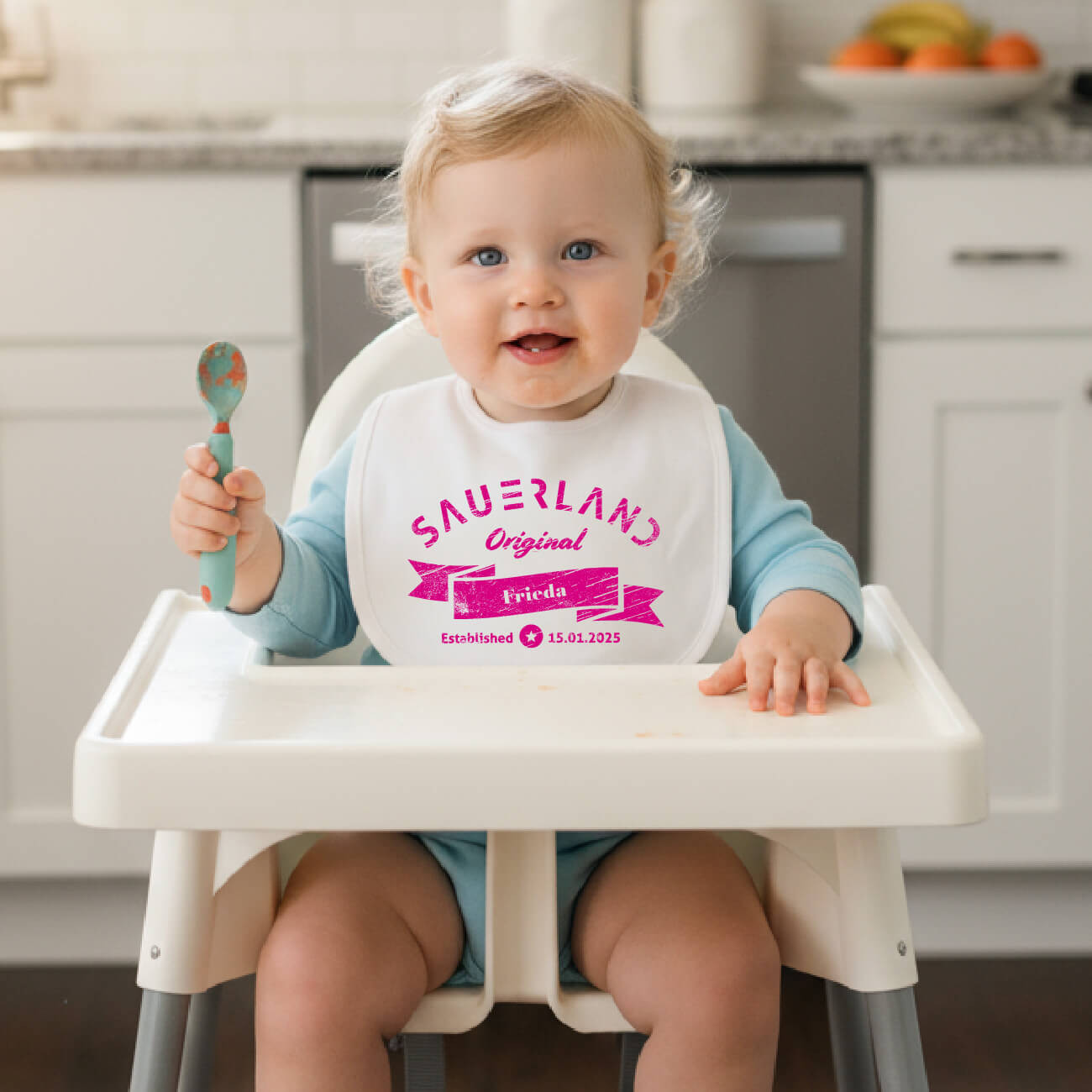 Kleines Mädchen sitzt in einem Kinderstuhl in der Küche und hat ein weißes Lätzchen mit dem Aufdruck Sauerland Original und dem Namen Frieda und ihr Geburtsdatum an.