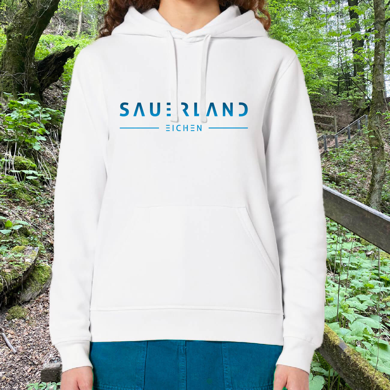 Frau in weißem Hoodie aus Biobaumwolle mit in einem blau-hellblau verlaufenden Aufdruck Sauerland Eichen steht auf einem Weg im Wald