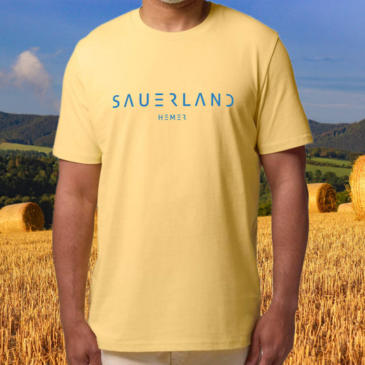 Mann trägt eine gelbes Sauerland T-Shirt mit blauem Aufdruck Sauerland Hemer und steht vor Strohballen. Im Hintergrund sind Sauerländer Hügel zu sehen.