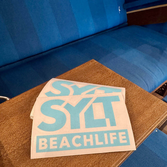 Sylt Aufkleber Beachlife in türkis liegt auf Ablagebrettchen eines Strandkorbes.