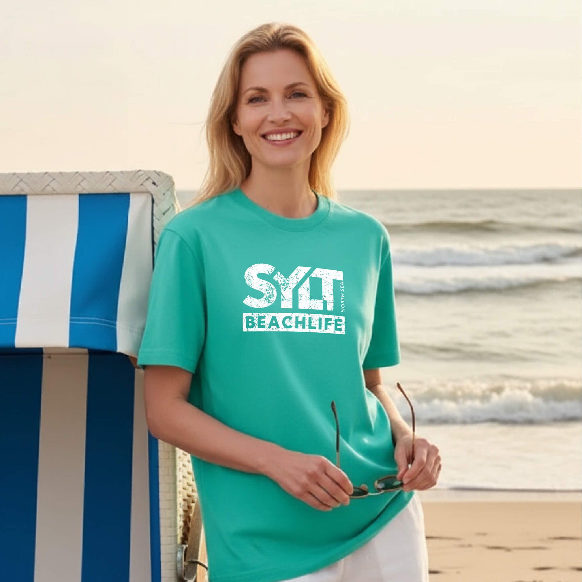 Frau trägt mint-grünes Sylt Beachlife T-Shirt aus Bio-Baumwolle, steht an einen Strandkorb gelehnt mit Nordsee im Hintergrund