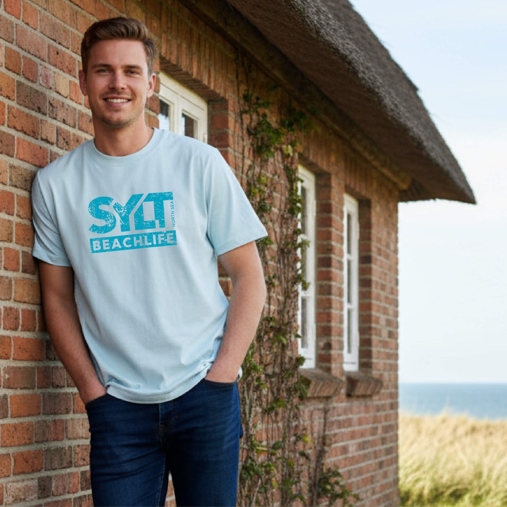 Mann in eisblauem Sylt Beachlife T-Shirt im Relaxed Fit steht an ein Friesenhaus angelehnt mit Nordsee im Hintergrund