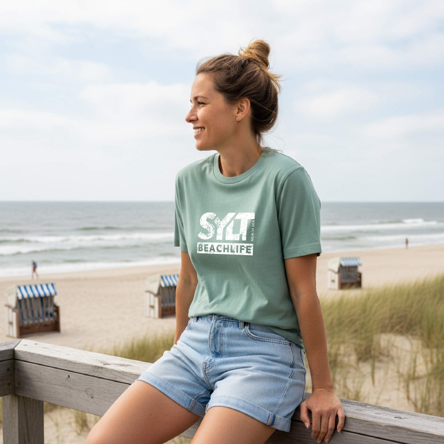 Frau in heidegrünem Sylt T-Shirt sitzt auf einem Holzgeländer. Im Hintergrund sieht man Dünen, Strandkörbe am Strand und die Nordsee.