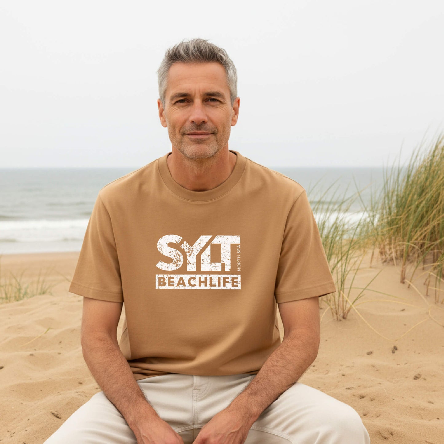 Mann trägt sandfarbenes Sylt Beachlife T-Shirt aus nachhaltiger Bio-Baumwolle und sitzt im Sand mit Dünen und Nordsee im Hintergrund