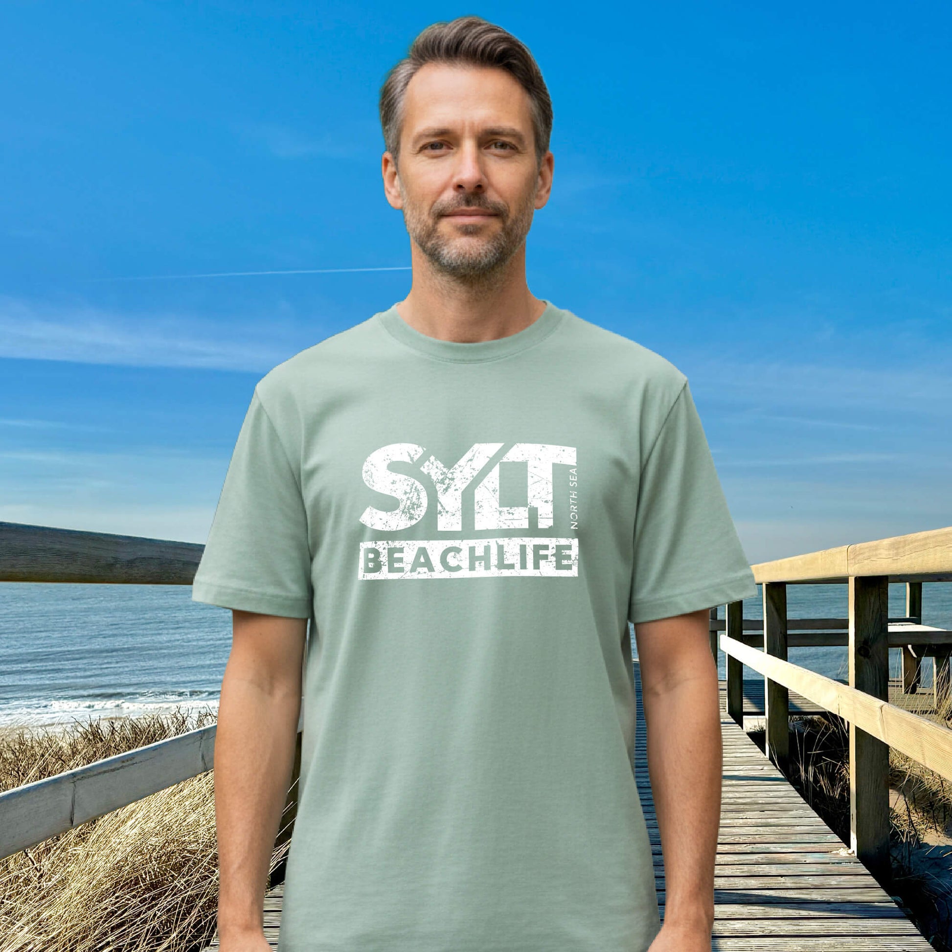 Mann in heidegrünem Sylt T-Shirt steht auf Holzsteg mit Meerblick und Dünenlandschaft im Hintergrund