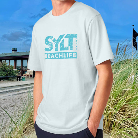 Mann in eisblauem Sylt Beachlife T-Shirt im Relaxed Fit vor typischem Nordsee-Strandhaus mit Dünen